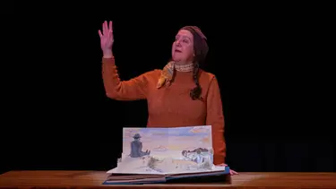 A woman in a orange sweater stands behind a table containing a fold out picture book. Her right arm is raised.