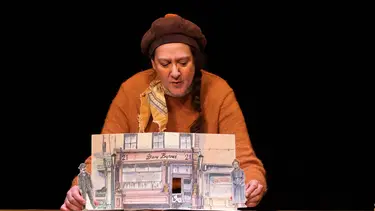 A woman leans over a fold out picture book as if displaying it to a crowd. The page shows and shop front with a man and woman on either side.