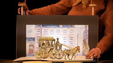 An image of a horse and carriage going down a street is displayed by a woman through a fold out picture book.