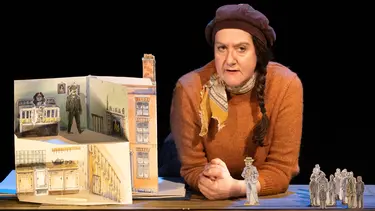 A woman in an orange sweater and brown hat leans over a table. On the table is an elaborate paper set of a home.