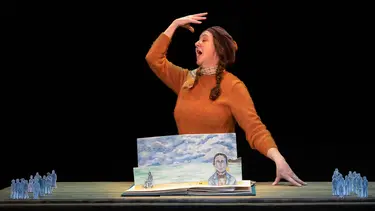 A woman sings with one arm raised towards her face and the other resting on a table. The table contains paper cut outs of illustrated people and a fold out picture book.