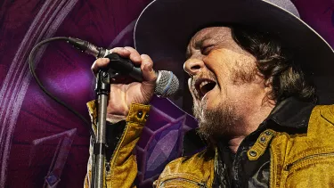 A man in yellow jacket and a cow boy hat singing in to the mic.