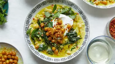 A white ceramic bowl with black ornate edge pattern holding spiced chickpea stew with coconut and turmeric on a tabletop. Five smaller bowls surround the edge of frame holding ingredients.