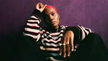 Arlo Parks wearing a striped knitted sweater, sitting on the ground against a dark purple wall.