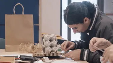 A student seated at a table working with mixed media including an egg carton, cardboard and string.