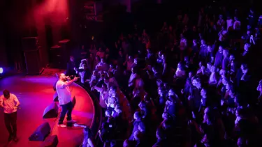 Briggs performing on stage under a red light, as viewed from the back and to one side of the theatre.