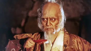 An elderly man with white hair and a long white beard holds a small red dish. He is wearing a robe.