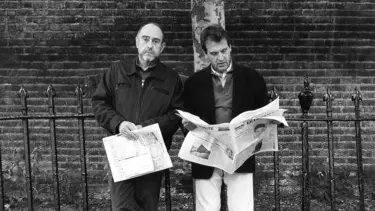 Two men with newspapers, standing in front of a brick wall and iron gates.