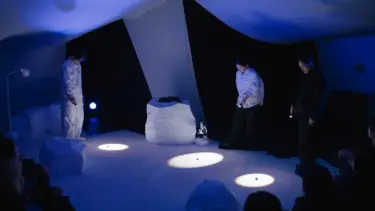 A full set that resembles a white tent structure is visible. Three cast members shine torches on objects that are on the floor.