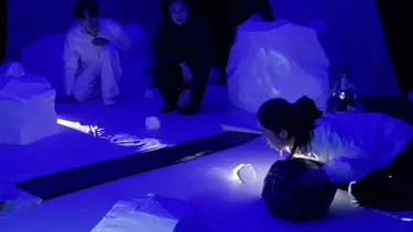 Three performers are crouched around a white coloured set. It is dimly lit and they are shining a light across a miniature river.