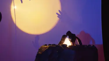 A penguin puppet is sitting on top of a black rock. A circular light with the shadows of peoples hands is visible in the background.