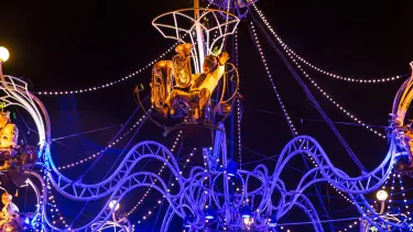 A large carousel decorated to resemble a chandelier is suspended in the air. Performers sit around the edges playing instruments.