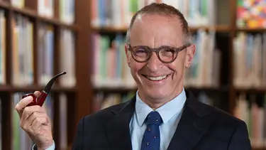 A man stands smiling in front of book shelves. He is wearing glasses, a suit and tie and is holding up a pipe.
