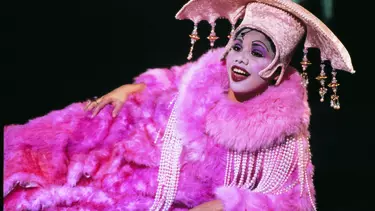 Deborah Mailman laying on her side in a bright pink fluffy coat draped with beads and wearing an embellished headpiece.