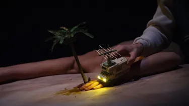 A mini toy van drives over a small snowy hill with its tiny headlights on. A hand is controlling it.