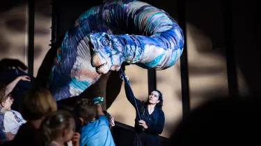 A performer manipulates a large, colorful dinosaur puppet on stage, captivating an audience of children with its lifelike movements.