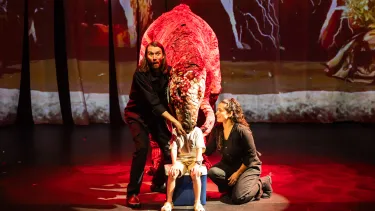 A child sits on stage with a dinosaur puppet head over them, guided by two performers, under dramatic red lighting.