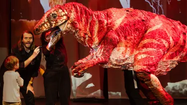 A child and an adult excitedly interact with a large, realistic red dinosaur puppet on stage, set against a dynamic backdrop.
