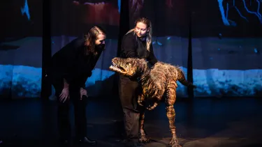 Two performers operate a realistic dinosaur puppet on a dimly lit stage with a colorful backdrop, creating a lifelike scene.