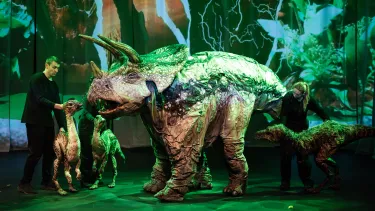 Puppeteers animate life-sized dinosaur puppets on stage, set against a lush, green backdrop, creating a realistic prehistoric scene.