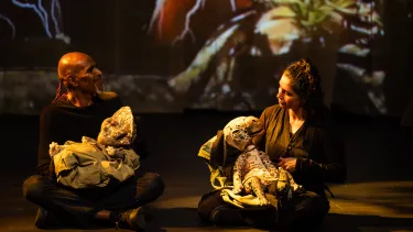 Two people sit cross-legged on stage, each holding a puppet, under warm lighting with a colorful backdrop.
