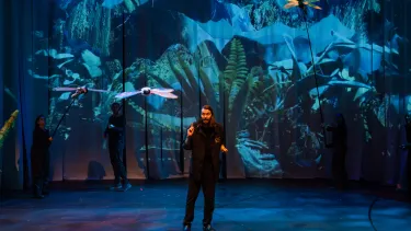 Performer on stage with dragonfly puppets, vivid jungle backdrop, and crew in black manipulating the scene.