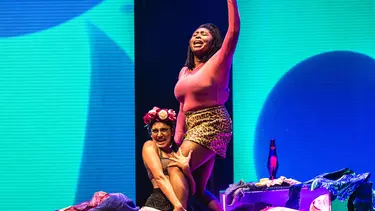 A woman holding up another woman in a pink top and leopard print skirt, on set with a lava lamp background.