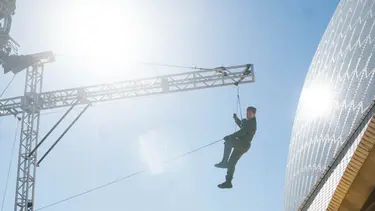 A man on a wire hangs off a crane next to the Sydney Opera House sails