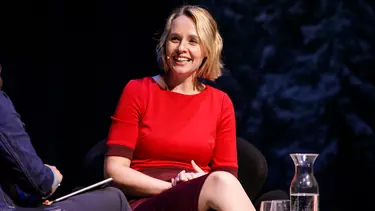 A person in a red t-shirt sitting on stage and smiling as if in conversation.