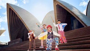 Four children in bright clothes pose on the steps of the Sydney Opera House on a bright sunny day.