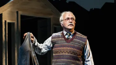 An older man with grey hair and a moustache stands wearing a wool vest, shirt and tier holding a blazer jacket. He is on a set that resembles a backyard at night.