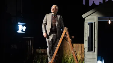 A man in a grey suit and maroon wool vest stands atop a ladder next to a shed.