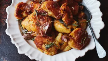 A pile of roasted potatoes on a white plate on a dark tabletop, with a silver spoon resting on the side of the plate.