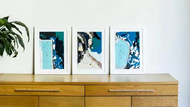 Three framed blue pictures resting on a wooden cabinet against a white wall.