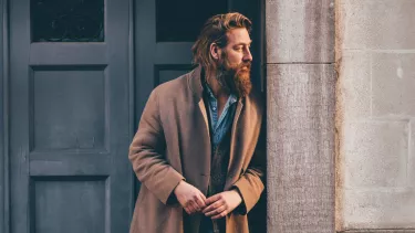 A man with brown hair and a long brown beard wearing a camel-colored coat over a denim shirt, leans on a door frame.