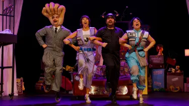 Four performers in whimsical costumes, including a man with a giant hand on his head and a man with bull horns, jump joyfully in a line on stage.