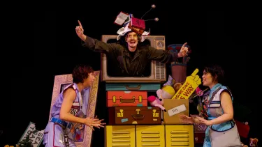 A man wearing a stack of books as a hat peers through a vintage television set at two women standing beside a colourful tower of suitcases.