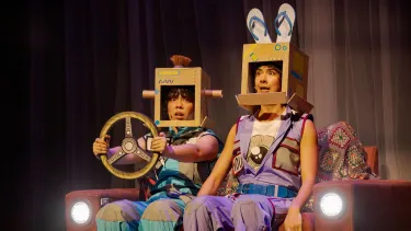 Two women wearing cardboard boxes on their heads—one topped with a brush and the other with blue thongs—sit on a red sofa while pretending to drive.