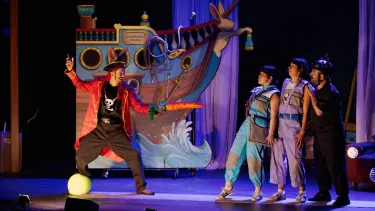 A pirate with rabbit ears stands on a giant tennis ball and points a large carrot towards three startled characters in front of a prop ship.