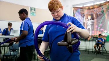 A boy with red hair holding a shaped balloon in a classroom.