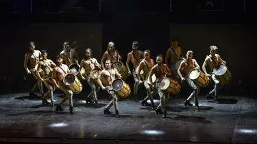 A group of 13 people playing drums on a stage.