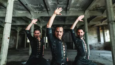 A group of three men in black clothes holding one hand above their heads in an empty warehouse.