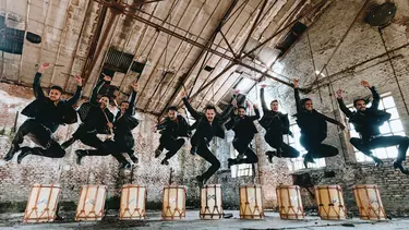 A group of nine people jumping in the air, with drums on the ground in an empty warehouse.