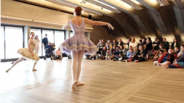 Dancers in dresses, dancing in front of a seated audience in a dance studio.