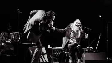 Nick Cave, a tall singer moves energetically during performance while Warren Ellis is seated next to him, singing into a microphone.
