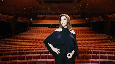 A woman in a black top, hand on her hip standing in front of an empty red theatre.