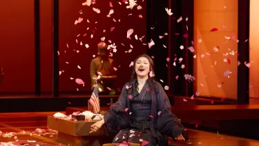 A woman kneels smiling next to a small US flag as petals fall down onto her.