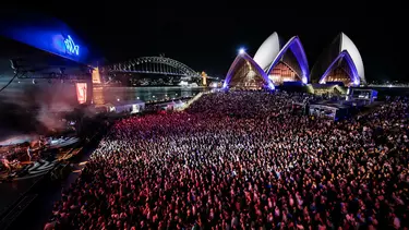 A large crowd of people watch a concert on the Opera House Forecourt at nighttime. The Sydney Harbour Bridge is in the background.