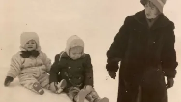 Two children sliding in the snow behind a young woman in a coat.