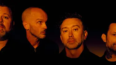 Four men stand side by side. They are all wearing black and are lit in an orange hue.